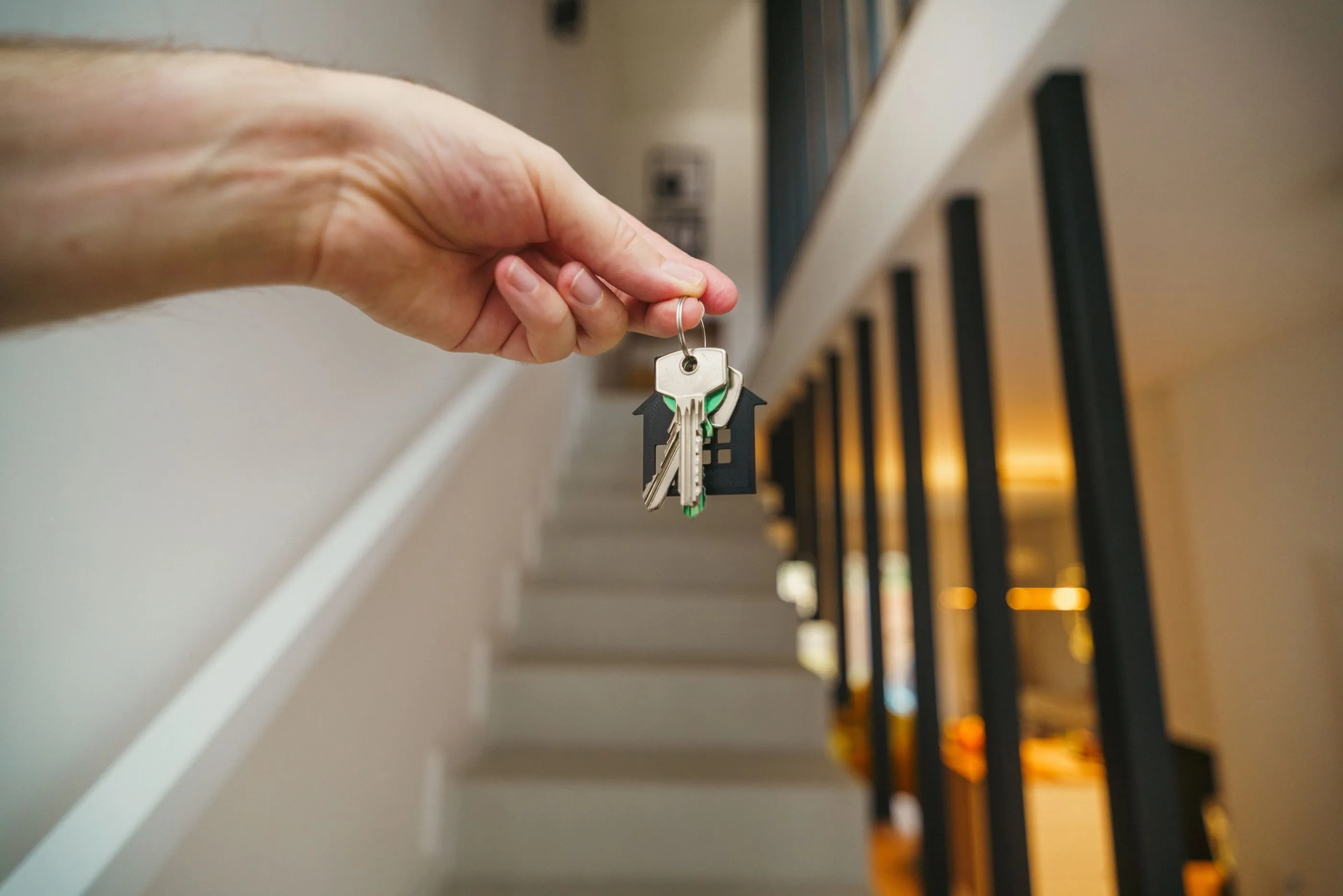 A hand holding a set of house keys on a bright staircase landing, warm light glowing from the room ahead.